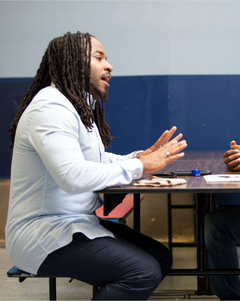 A mentor sits across the table from two young men, listening with open hands